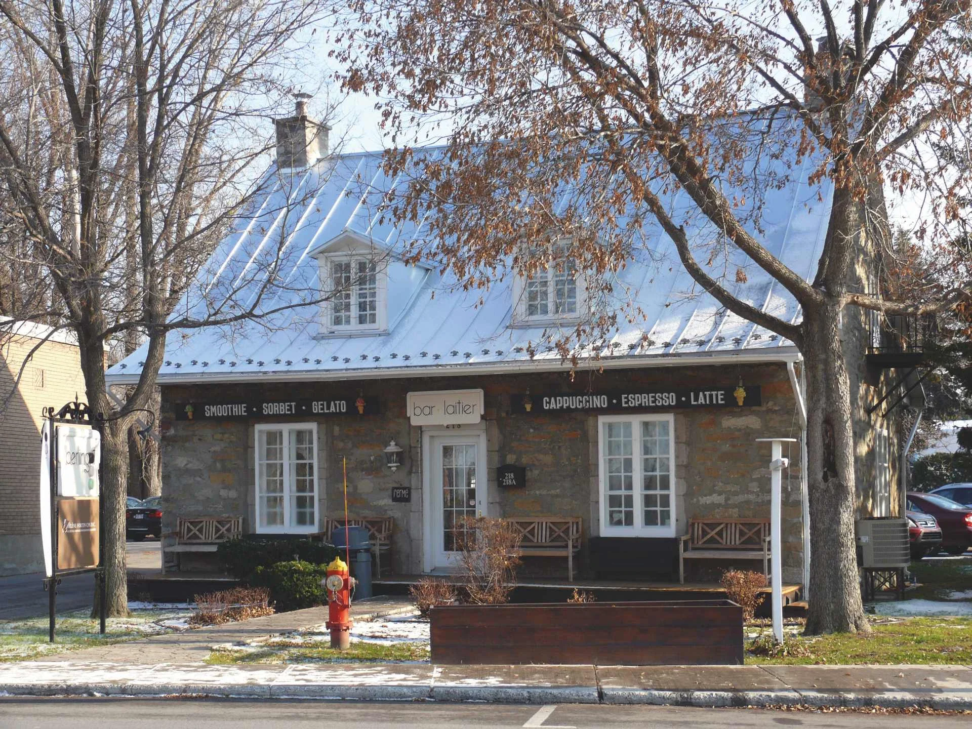 Maison de pierre ancienne au toit de tôle claire, transformée en bar laitier ; enseignes « smoothie, sorbet, gelato » sous l'auvent, bancs de bois et arbres dénudés devant.