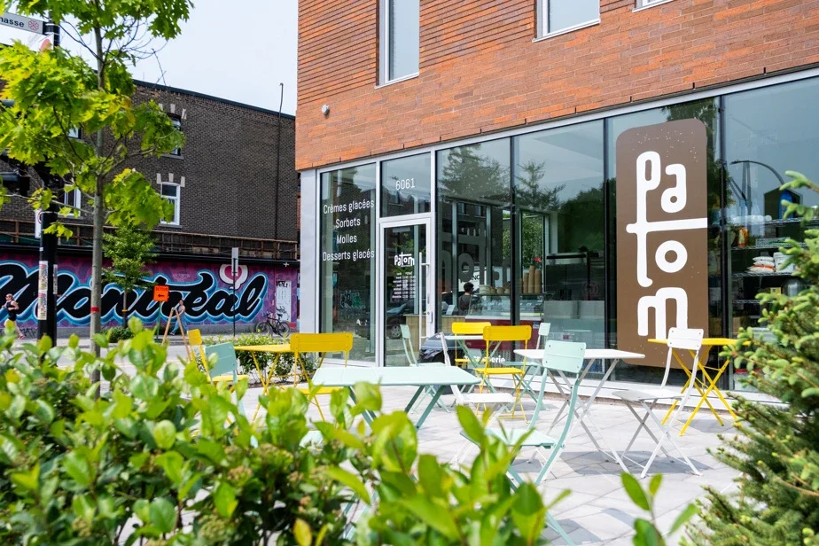 Devanture vitrée de la crèmerie, enseigne « Patom » à la verticale ; terrasse de chaises et de tables de bistro jaunes et vert d'eau sur le trottoir.